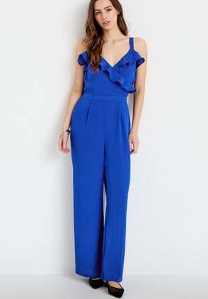 Frau trägt einen kobaltblauen ärmellosen Overall mit gerafftem V-Ausschnitt und weitem Bein, steht vor einem schlichten weißen Hintergrund.