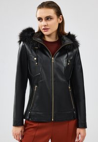 Giacca nera in pelle con cappuccio in pelliccia sintetica. Presenta una chiusura con zip sul davanti, tasche sul petto inclinate e dettagli dorati. Texture liscia e vestibilità su misura.