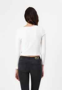 Top largo de manga larga blanco con textura acanalada, combinado con jeans ajustados negros que presentan un logo dorado en la cinturilla y bolsillos traseros destacados.