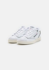 Bílé kožené tenisky s hladkou texturou, s klasickým šněrovacím designem a brandingem Reebok na jazyku a na boku.
