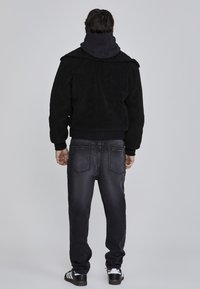 Chaqueta de forro polar negra con puños y cuello acanalados, usada sobre una sudadera con capucha negra. Combinada con jeans de mezclilla oscura y zapatos negros.