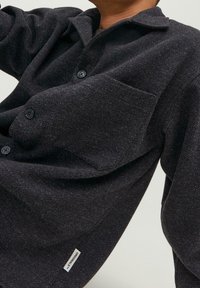 Camicetta nera con bottoni in tessuto testurizzato, dotata di tasca sul petto e vestibilità rilassata. Include un piccolo etichetta del marchio vicino all'orlo.