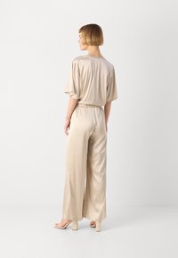 Beige satin jumpsuit with relaxed fit, short sleeves, elastic waist, and wide legs, paired with light-colored heeled sandals.