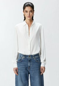 Camicia bianca a maniche lunghe con colletto classico. Indossata con jeans a vita alta larghi di un blu chiaro con dettagli dorati.