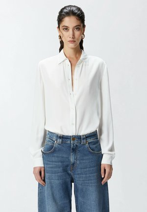 Camicia bianca a maniche lunghe con colletto classico. Indossata con jeans a vita alta larghi di un blu chiaro con dettagli dorati.