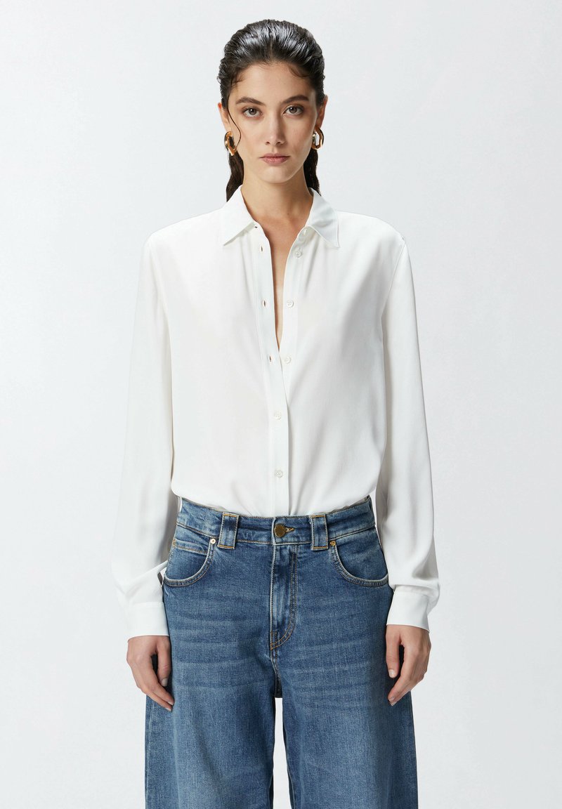Camicia bianca a maniche lunghe con colletto classico. Indossata con jeans a vita alta larghi di un blu chiaro con dettagli dorati.