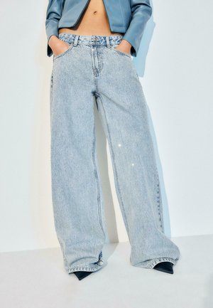 Person iført lyseblå vidde jeans og en kort blå jakke, hænderne stukket i lommerne, mørke spidse sko synlige under jeansene.