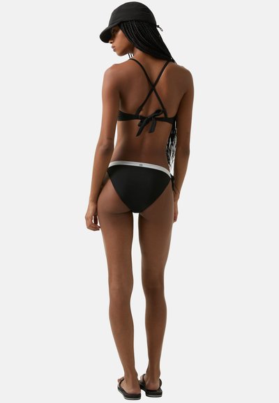 Černý bikini s překříženým designem ramínek, zavazováním vzadu a minimálním zakrytím. Kalhotky s vysokým pasem mají bílý akcentní pás.