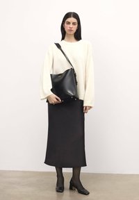 Borsa a tracolla in pelle nera con una forma curva, hardware dorato e cinque chiodini sul fondo; indossata con un maglione ampio color crema e una gonna midi nera.