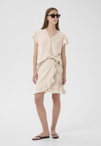 Bežs lina tops ar īsām piedurknēm un pogām, apvienots ar atbilstošu apņemšanas svārku ar frill apakšmalu. Valkāts ar melnām flip-flop sandalēm.