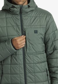 Green puffer jacket with a quilted texture, zip front, adjustable hood, and a small chest pocket with a logo patch.