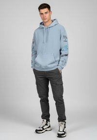 Ljusblå huvtröja med grafiska tryck på ärmarna, framficka i kängurustil, i kombination med mörka cargobyxor och svart-vita sneakers.