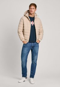Chaqueta acolchada beige con capucha, con un diseño de costura horizontal, combinada con una camiseta azul marino y vaqueros azules, complementada con zapatillas blancas.