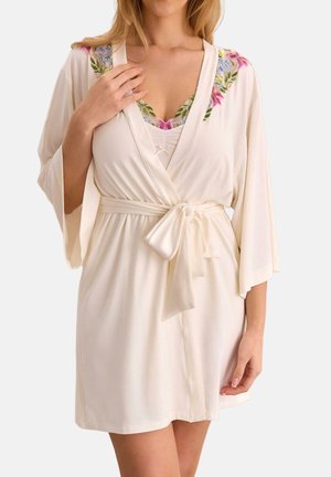 Kobieta ubrana w kremowy krótki szlafrok z przewiązanym paskiem w talii i kwiatowym haftem na ramionach, nosi dopasowany top z kwiatowym wykończeniem.