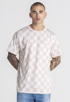 Man met tatoeages draagt een beige en wit geruit overhemd met korte mouwen en een blauwe spijkerbroek, staand tegen een effen achtergrond.