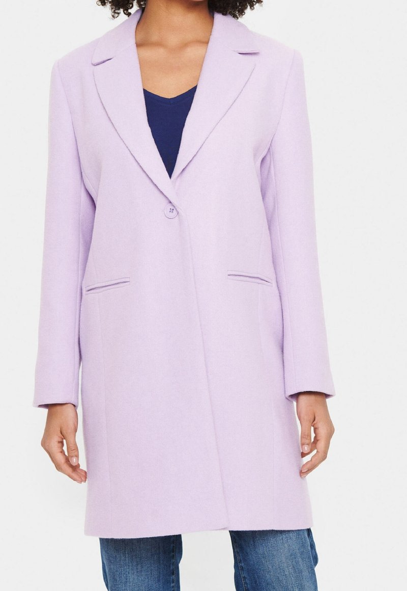 Cappotto in misto lana lilla con chiusura a un bottone, rever a punta, due tasche frontali e taglio dritto. Indossato sopra un top blu navy.