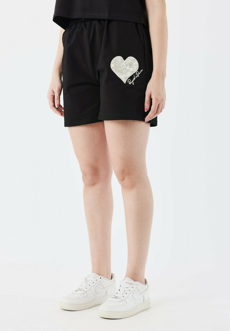Shorts noirs avec une taille élastique présentent un grand motif en forme de cœur blanc et du texte. Portés avec des baskets blanches. Matière en coton lisse.