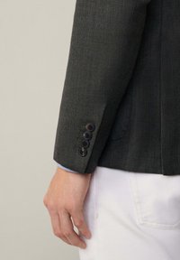 Braccio destro in un blazer scuro e texturizzato con quattro bottoni neri sul polsino, un polsino di camicia blu visibile e pantaloni bianchi con tasca posteriore.