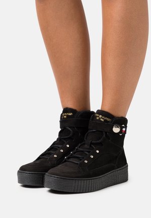 Baskets montantes en daim noir avec une semelle plateforme texturée, des œillets métalliques et une bride ornée d'un logo et d'un accent tricolore.