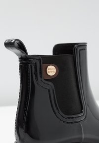 Musta Chelsea-saapas kiiltävällä viimeistelyllä, joustavilla sivuilla ja pronssinvärisellä logoyksityiskohdalla. Sileä pinta saumoilla korostettuna.