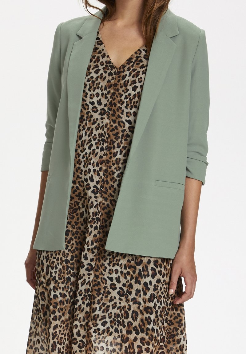 Kvinde iført en lys grøn blazer over en V-hals leopardmønstret kjole, med ærmerne på blazeren afslappet optrukket.