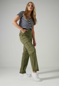 Groene cargobroek met meerdere zakken en een relaxte pasvorm, gecombineerd met een marineblauw en wit gestreept shirt met korte mouwen en witte sneakers.