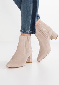 Botas de ante beige hasta el tobillo con punta afilada, paneles laterales texturizados y un tacón bajo en bloque. Llevadas con jeans oscuros de mezclilla con vuelta.