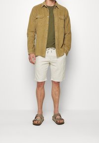 Hnědá kordurová košile se dvěma náprsními kapsami, olivově zelené tričko, krémové šortky a hnědé sandály. Neformální outfit s vrstvenými texturami.