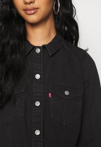 Frau trägt ein schwarzes Hemd mit aufgesetzten Brusttaschen und silbernen Druckknöpfen, Creolen und lockigem, offenem Haar.