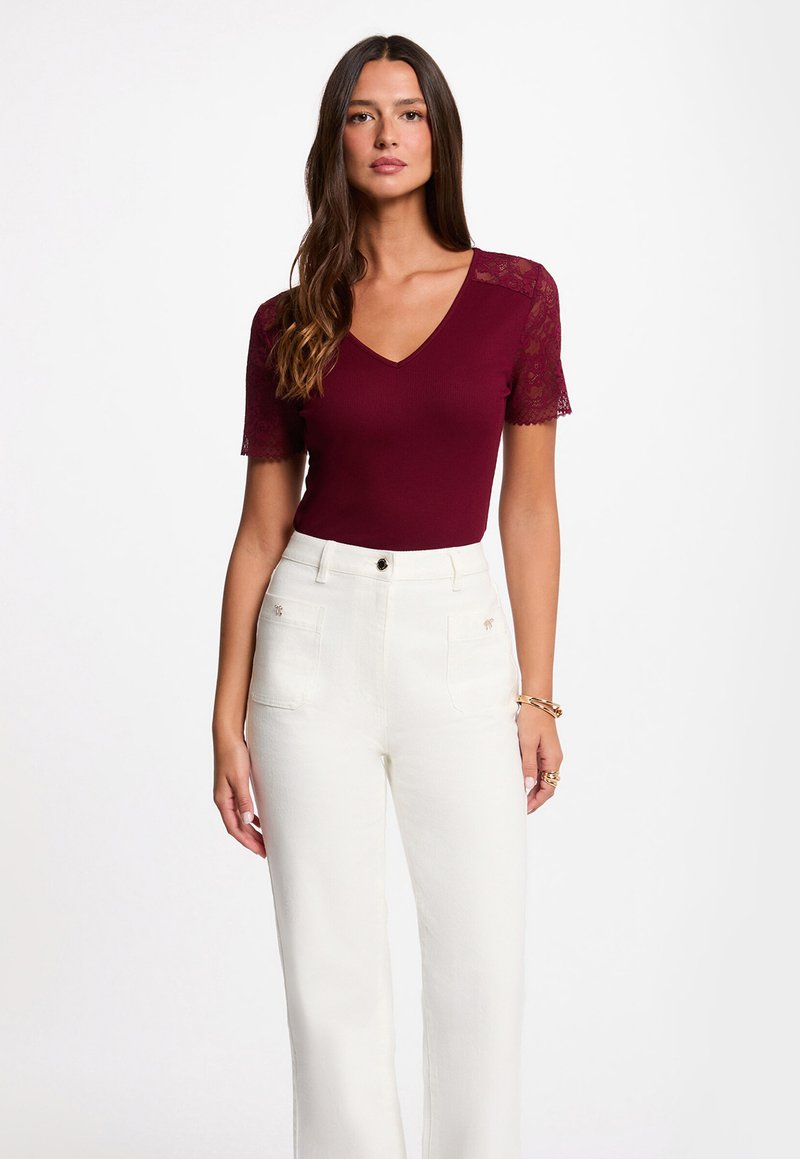 Woman wearing a burgundy V-neck top with lace sleeves and high-waisted white pants, standing against a plain white background.