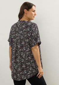 Donna che indossa una camicia nera ampia con motivi floreali bianchi e viola, in piedi di lato contro un muro chiaro e semplice.