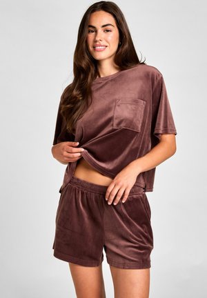 Femme portant un haut marron doux à manches courtes avec poche et un short assorti à taille élastique, souriante et relevant légèrement le haut.