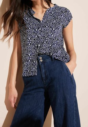 Kvinde iført en kortærmet navybluse med hvide geometriske mønstre og høj taljede mørkeblå bukser med vide ben og to knapper.