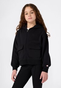 Niña joven con pelo largo y rizado que lleva una sudadera con cremallera negra y dos bolsillos frontales, así como pantalones negros a juego, de pie contra un fondo blanco.