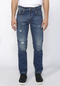 Jeans de mezclilla azul con un ajuste Slim, que presentan desgastes, parches rasgados y un diseño clásico de cinco bolsillos. Combinados con zapatillas negras.