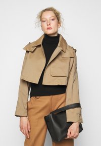 Beskjært beige jakke med bred krage, frontlomme og knappe detaljer, brukt over en svart genser med høy hals og parret med brune bukser og en svart veske.