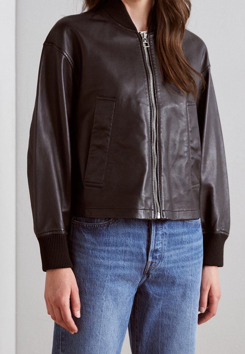 Femme portant une veste en cuir brun foncé à fermeture éclair avec des poignets côtelés et un jean bleu clair, debout devant un fond uni.