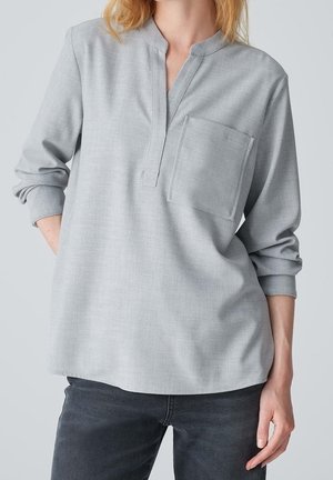 Femme portant une chemise à manches longues gris clair avec une poche poitrine et un jean gris foncé sur un fond uni.