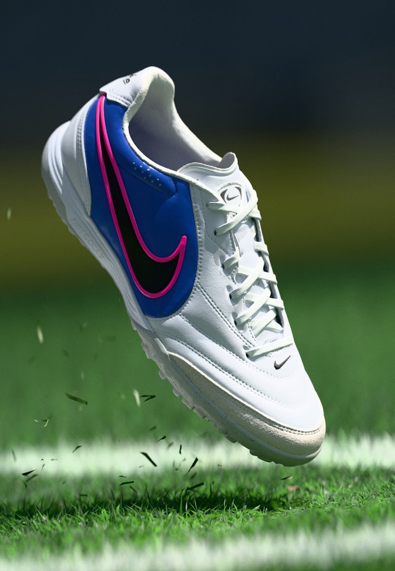 Λευκό και μπλε ποδοσφαιρικό παπούτσι Nike με ροζ και μαύρο σήμα, που πετάει χόρτο σε έναν πράσινο αγωνιστικό χώρο κοντά σε μια λευκή γραμμή.