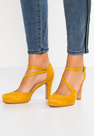 Fødder iført sennepsgule ruskindshøjhælede sko med ankelsnører, kombineret med blå cropped jeans med sidelommer.