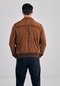 Man die een bruine suède jas en marineblauwe broek draagt, met zijn rug naar voren en zijn handen in de zakken, staande tegen een effen achtergrond.