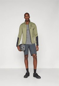 Olivengrønn jakke med svarte detaljer, grå t-skjorte, grå shorts, holder en grå hjelm, står foran en nøytral bakgrunn.