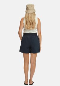 Námořnické kraťasy s elastickým pasem v kombinaci s bílým tílkem a béžovým kloboukem; dlouhé, splývavé vlasy jsou viditelné.