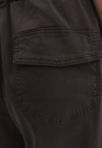 Donkergekleurde cargobroek met een grote, rechthoekige zak op de achterkant, met zichtbare stiksels en een lichte glans op het materiaal.