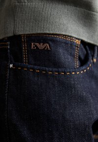 Mörka jeans i denim med orangea sömmar, som har en subtil textur. En bronsfärgad logotypmärke med texten "EVA" syns på bakfickan.