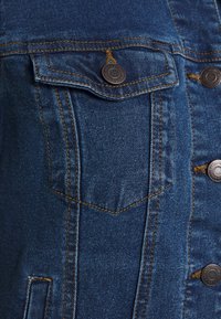 Detalj av en jeansjacka som har en bröstficka, bruna sömmar, metallknappar och ett texturerat blått tyg.