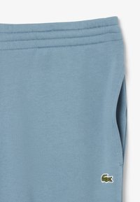 Vaatekankaalta pehmeät vaaleansiniset verkkarit, joissa on joustava vyötärö ja pieni vihreä krokotiililogo alhaalla vasemmalla puolella.