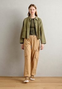 Olivgrüne Jacke mit Fronttaschen, braunes Rippoberteil, beigefarbene Cargo-Hosen mit Seitentaschen und hellbraune Sneakers. Hintergrund aus Holzfußboden.