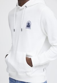Haut à capuche en polaire blanc avec une poche avant, des cordons de serrage et un logo navy représentant un soleil et un palmier. Tissu texturé avec des poignets côtelés.