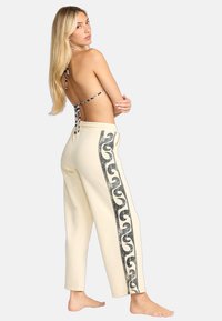 Pantalones jogger crema con un patrón de espiral de purpurina negra a los lados. Modelo vistiendo un top halter de patrón en blanco y negro, descalza, vista desde atrás.